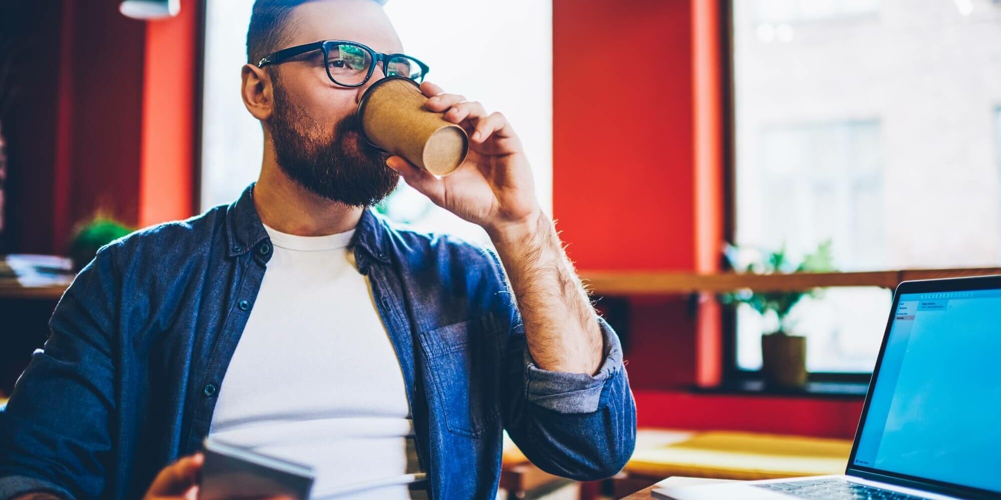 Bearded male IT developer with smartphone in hands drinking tasty coffee to go while working remotely at modern laptop connected to 4G internet.Young man blogger enjoying beverage while installing app